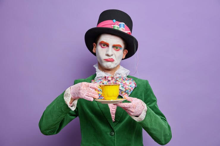Foto gratuito annoiato dispiaciuto cappellaio uomo ossessionato dal bere il tè indossa un grande cappello verde costume papillon posa contro il muro viola arriva su una festa spettrale celebra halloween stand al coperto