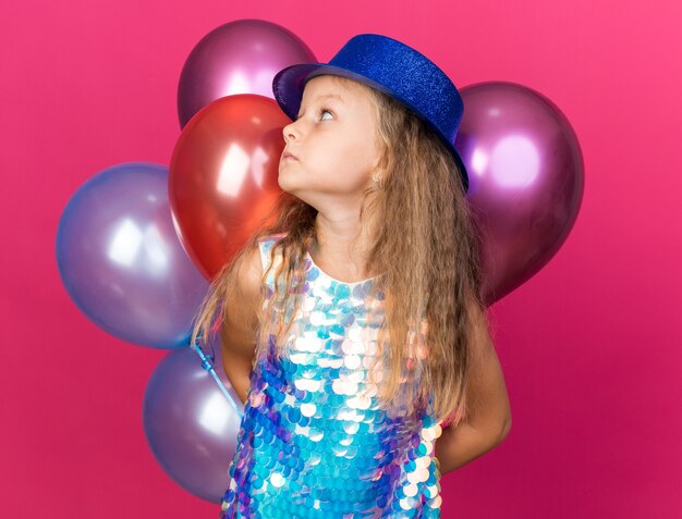 ansiosa bambina bionda con cappello da festa blu che tiene palloncini di elio e guarda il lato isolato sulla parete rosa con spazio per le copie