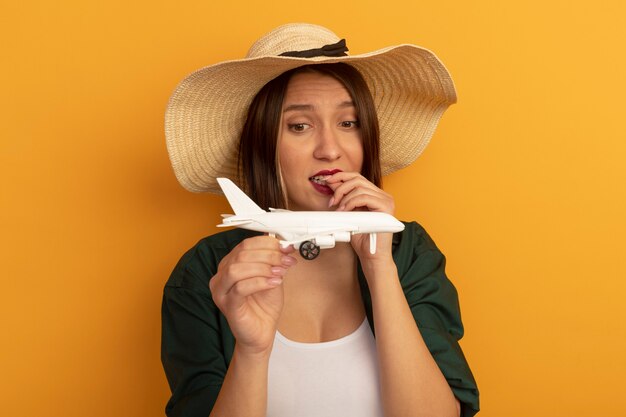 Ansiosa donna graziosa con cappello da spiaggia morde le unghie tenendo e guardando il modello aereo isolato sulla parete arancione