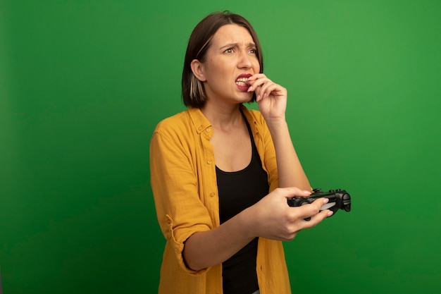 Ansiosa donna graziosa morde il chiodo e tiene il controller guardando il lato isolato sulla parete verde