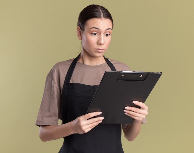 Ansiosa giovane barbiere bruna in uniforme che tiene e guarda gli appunti clip