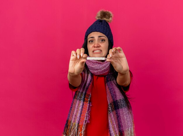 Ansiosa giovane donna malata che indossa cappello invernale e sciarpa che allunga il termometro verso la parte anteriore guardando davanti isolato sulla parete rosa con lo spazio della copia