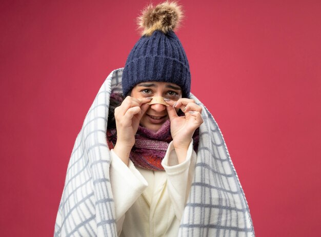 Ansiosa giovane donna malata che indossa un abito invernale cappello e sciarpa avvolti in un plaid guardando davanti mettendo intonaco sul naso isolato sul muro rosa