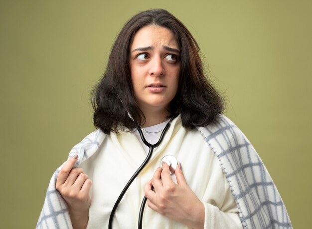 Ansiosa giovane donna malata che indossa veste e stetoscopio avvolto in un plaid ascoltando il suo battito cardiaco afferrando il plaid guardando il lato isolato sul muro verde oliva