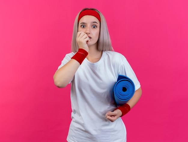 Ansiosa giovane donna sportiva con bretelle che indossano fascia e braccialetti morde le unghie e tiene il tappetino sportivo isolato sul muro rosa