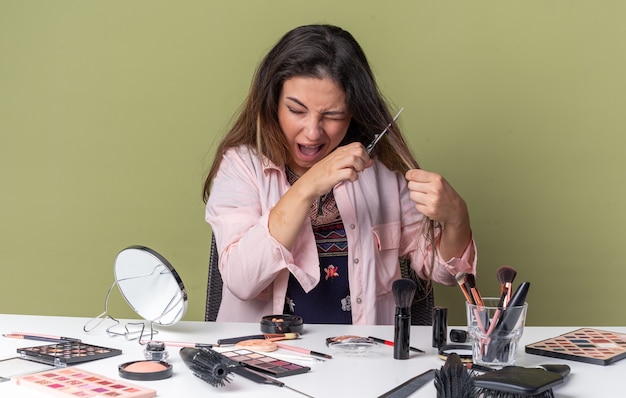 Ansiosa giovane ragazza bruna seduta al tavolo con strumenti per il trucco che si taglia i capelli con le forbici