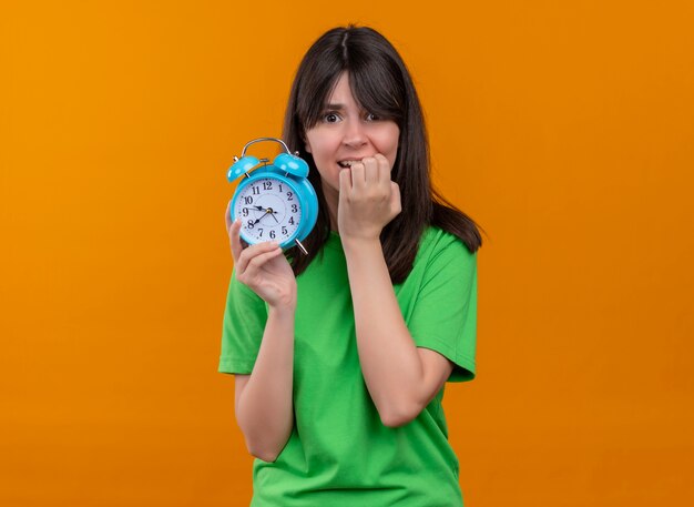 Ansiosa giovane ragazza caucasica in camicia verde tiene l'orologio e mette la mano sulla bocca su sfondo arancione isolato