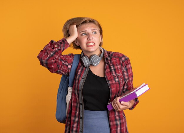 Ansiosa giovane studentessa slava con le cuffie che indossa lo zaino mette la mano sulla testa tiene libro e taccuino