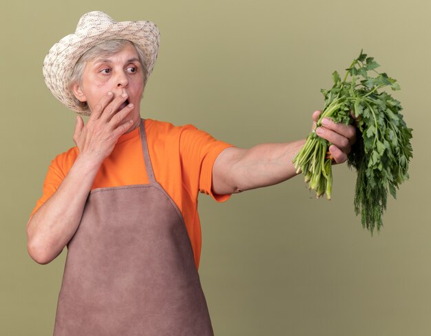Ansioso giardiniere femmina anziana che indossa cappello da giardinaggio mette la mano sulla bocca e tiene il mazzo di aneto di coriandolo su verde oliva