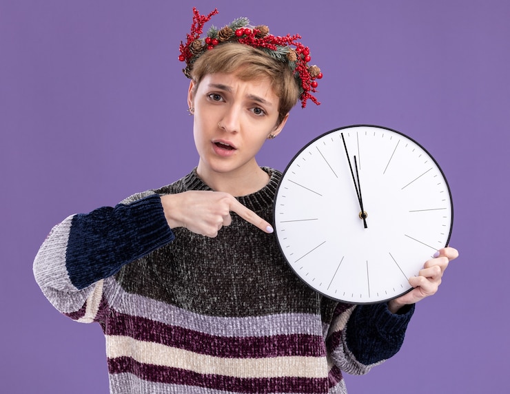 Foto gratuito ansioso giovane bella ragazza che indossa la corona di natale testa tenendo e indicando l'orologio isolato sulla parete viola