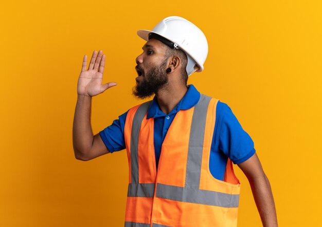 Ansioso giovane costruttore uomo in uniforme con casco di sicurezza che chiama qualcuno che guarda il lato isolato sulla parete arancione con spazio copia