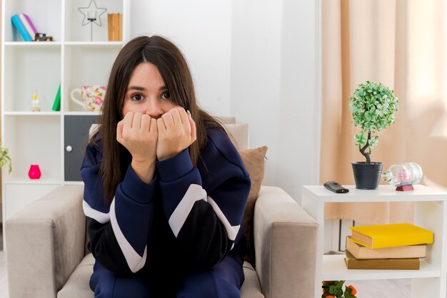 Ansioso giovane donna abbastanza caucasica seduto sulla poltrona nel soggiorno progettato mantenendo i pugni sulla bocca alla ricerca