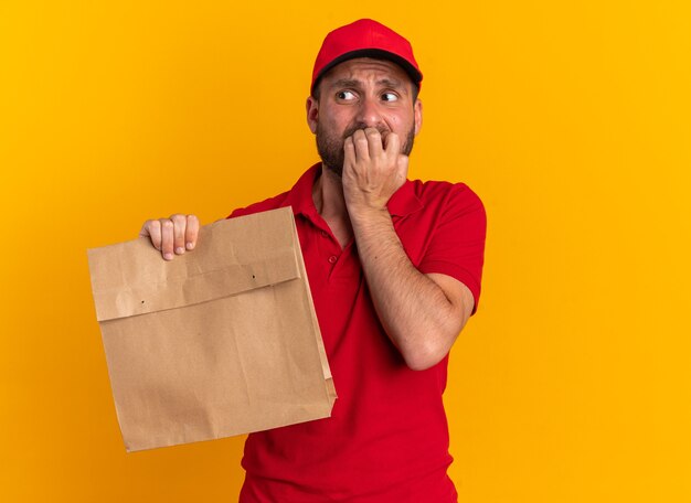 Ansioso giovane fattorino caucasico in uniforme rossa e cappuccio che tiene un pacchetto di carta tenendo le dita sul labbro guardando di lato