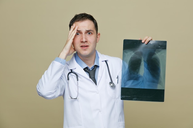 Ansioso giovane medico maschio che indossa una veste medica e uno stetoscopio intorno al collo che mostra un'inquadratura a raggi x tenendo la mano sulla testa guardando la fotocamera isolata su sfondo verde oliva