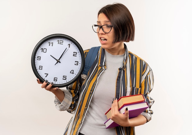 Ansioso giovane studente ragazza con gli occhiali e borsa posteriore in possesso di libri e orologio guardando l'orologio isolato su bianco