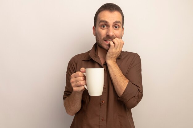 Ansioso giovane uomo caucasico tenendo la tazza di tè mettendo la mano sul labbro guardando la telecamera isolata su sfondo bianco con spazio di copia