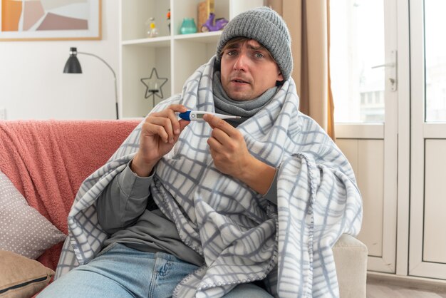 Ansioso giovane uomo malato con sciarpa intorno al collo indossando cappello invernale avvolto in plaid tenendo il termometro seduto sul divano in soggiorno