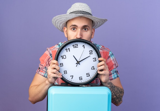 Ansioso giovane viaggiatore uomo con cappello da spiaggia di paglia che tiene l'orologio in piedi dietro la valigia isolata sul muro viola con spazio copia