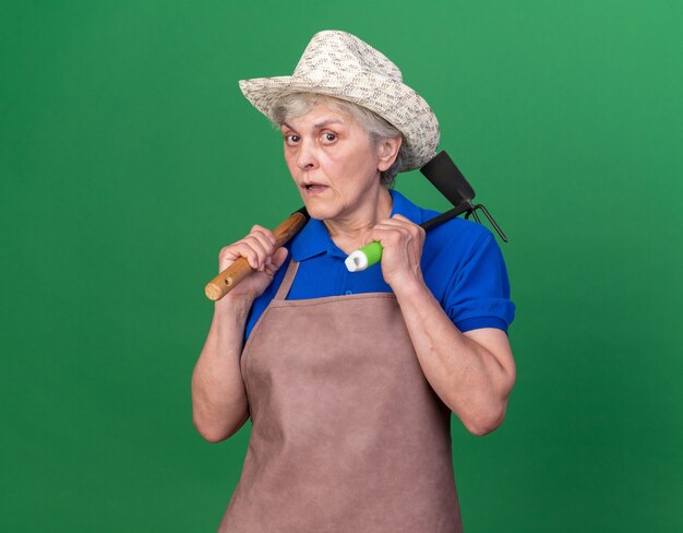Anziana giardiniera femmina ansiosa che indossa cappello da giardinaggio tenendo il rastrello e il rastrello della zappa sulle spalle