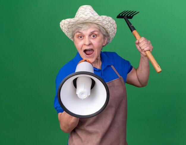 Anziana giardiniera femmina ansiosa che indossa cappello da giardinaggio tenendo rastrello e gridando in un altoparlante isolato sulla parete verde con spazio copia