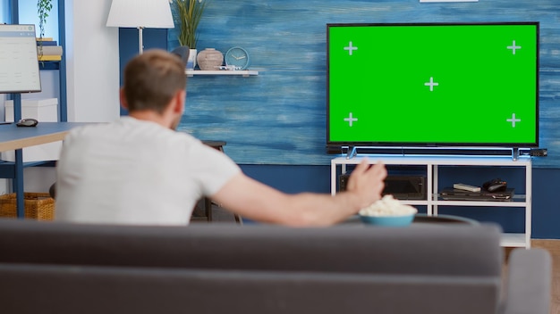 Appassionato di sport che guarda il gioco sul mockup della tv a schermo verde che incoraggia la squadra preferita mentre si rilassa a casa seduto sul divano. Sostenitore sportivo uomo che guarda la televisione con display chroma key in soggiorno.