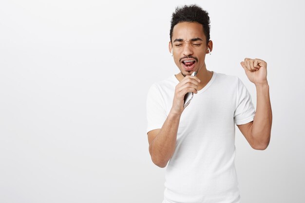 Appassionato ragazzo afro-americano che suona il karaoke, canta nel microfono dello smartphone, indossa le cuffie wireless