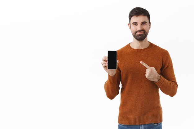 Applicazione tecnologica e concetto online Ritratto di un bell'uomo barbuto sicuro di sé consiglia di acquistare un gioco di download in abbonamento o un'app indicando il display del telefono cellulare e sorridendo soddisfatto