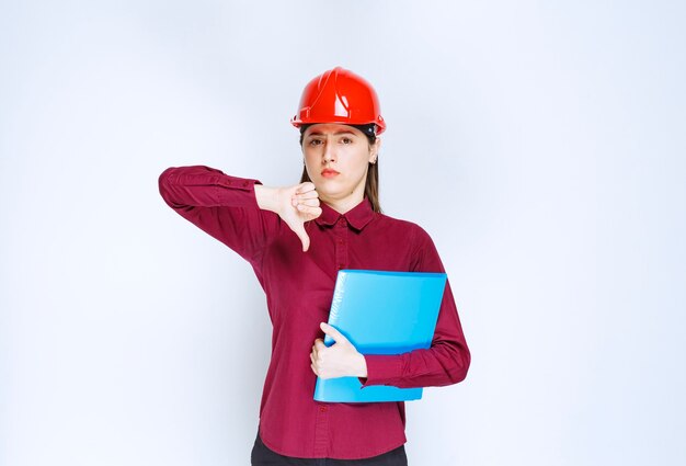 Architetto femminile in casco rosso che tiene cartella blu con documenti e dà i pollici in giù.