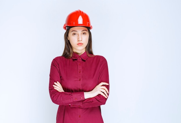 Architetto femminile sicuro in casco duro rosso che sta e che posa sopra la parete bianca.