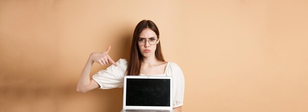 Arrabbiata donna accigliata con gli occhiali che punta allo schermo del laptop lamentandosi e sembra delusa in piedi