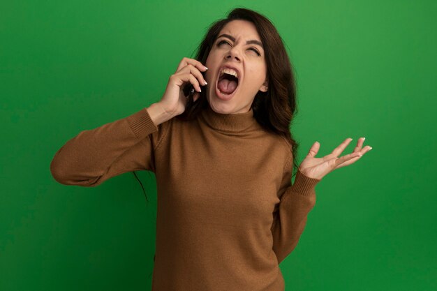 Arrabbiato alzando lo sguardo giovane bella ragazza parla sul telefono e diffondendo la mano isolata sulla parete verde