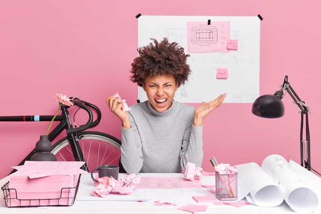 Arrabbiato architetto donna posa a tavola con progetti di carta e scarti arrabbiato per trovare l'errore nel suo lavoro di progetto esclama con espressioni indignate pose contro il muro rosa nello spazio di coworking