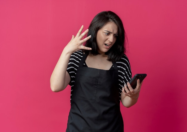 Arrabbiato giovane bella donna parrucchiere in grembiule guardando lo schermo del suo telefono cellulare con espressione infastidita sul rosa