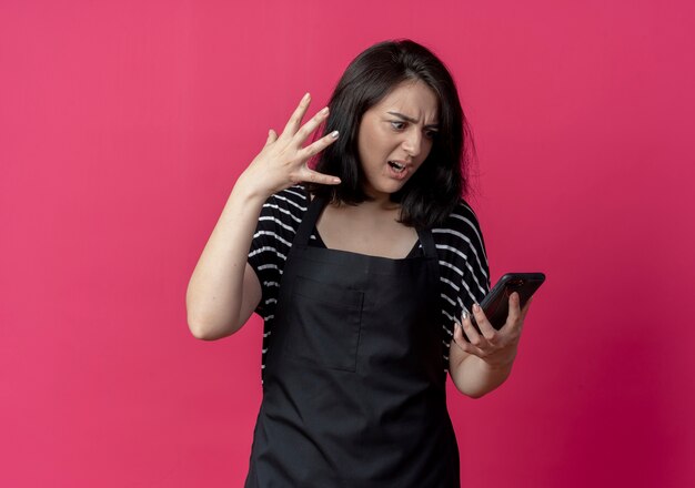 Arrabbiato giovane bella donna parrucchiere in grembiule guardando lo schermo del suo telefono cellulare con espressione infastidita sul rosa