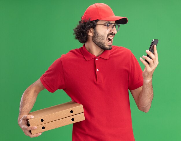 Arrabbiato giovane fattorino caucasico in uniforme rossa e berretto con gli occhiali che tengono pacchi di pizza e telefono cellulare guardando il telefono