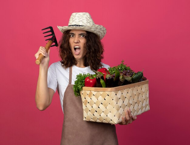 Arrabbiato giovane giardiniere femminile in uniforme che indossa il cappello da giardinaggio tenendo il cesto di verdure e sollevando rastrello isolato sul rosa