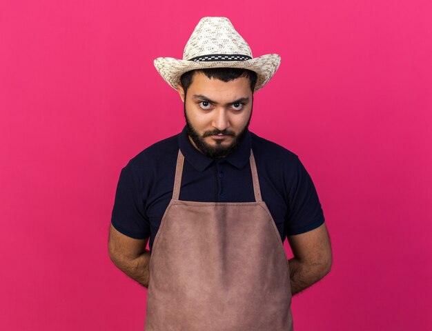 arrabbiato giovane giardiniere maschio caucasico indossando il cappello da giardinaggio tenendo le mani dietro e isolato sul muro rosa con spazio di copia