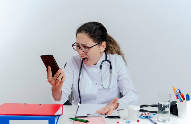 Arrabbiato giovane medico femminile che indossa abito medico e stetoscopio e occhiali seduto alla scrivania con strumenti medici che tengono guardando il telefono cellulare isolato