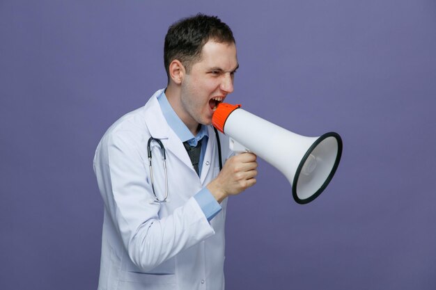 Arrabbiato giovane medico maschio che indossa accappatoio medico e stetoscopio intorno al collo in piedi nella vista di profilo guardando il lato parlando in altoparlante isolato su sfondo viola