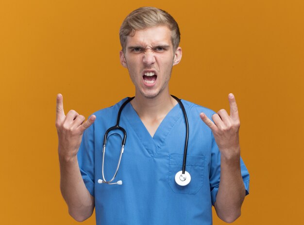 Arrabbiato giovane medico maschio indossa uniforme medico con lo stetoscopio che mostra gesto di capra isolato sulla parete arancione