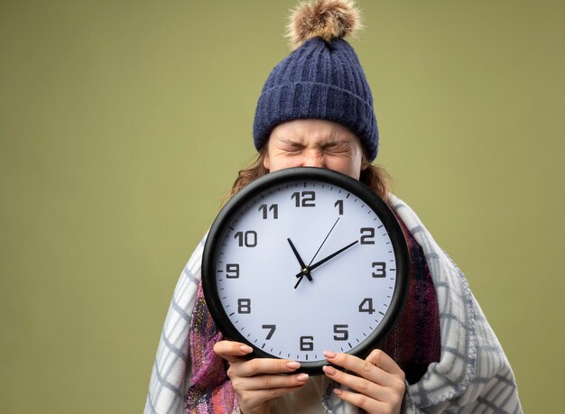 Arrabbiato giovane ragazza malata che indossa una veste bianca e cappello invernale con sciarpa viso coperto con orologio da parete avvolto in un plaid isolato su verde oliva