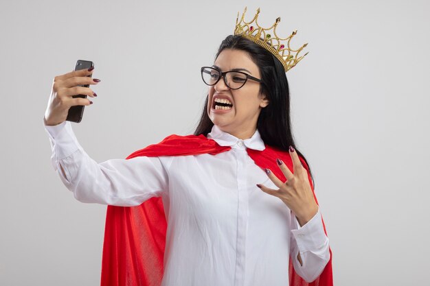 Arrabbiato giovane supereroe caucasico ragazza con gli occhiali e corona tenendo la mano in aria prendendo selfie isolati su sfondo bianco con copia spazio