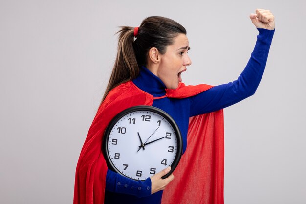 Arrabbiato giovane supereroe ragazza in piedi in vista di profilo tenendo l'orologio da parete e alzando la mano isolati su sfondo bianco