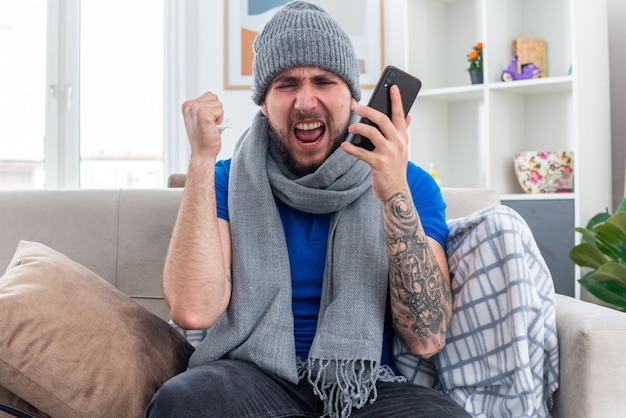 Arrabbiato giovane uomo malato che indossa una sciarpa e cappello invernale seduto sul divano in soggiorno tenendo tovagliolo e telefono cellulare guardando verso il basso il pugno di serraggio urlando