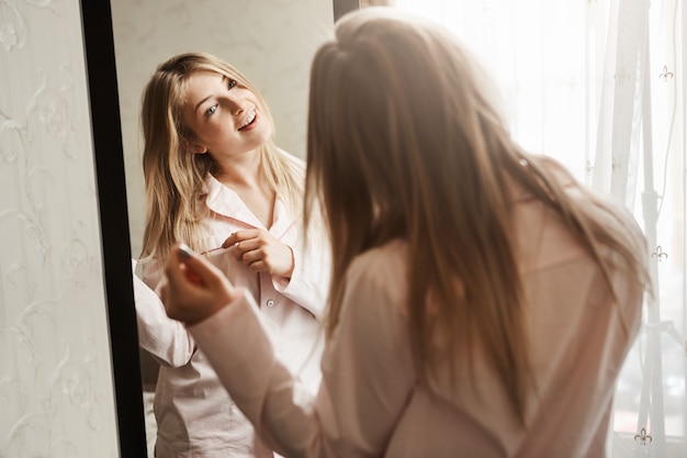 È arrivato il momento di vestirsi e di incontrare avventure. Colpo domestico di bella ragazza caucasica bionda che guarda in specchio, indossando indumenti da notte e toccando la ciocca di capelli, pensando alla nuova acconciatura