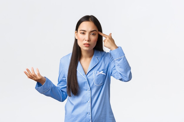 Arrogante ragazza asiatica che sembra frustrata e perplessa, indossa un pigiama blu, guarda con disprezzo mentre tocca tample e alza la mano confusa, rimproverando qualcuno che si comporta da stupido o pazzo