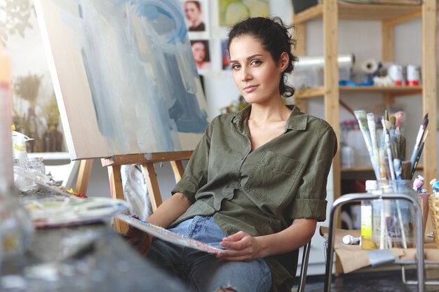 Artista di giovane donna caucasica di talento positivo dall'aspetto alla moda che si rilassa sulla sedia accanto al cavalletto in officina dopo aver finito di dipingere. Professione, creatività, arte moderna, lavoro e occupazione