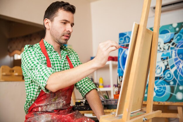 Artista maschio bello che indossa un grembiule e lavora a un dipinto nel suo studio