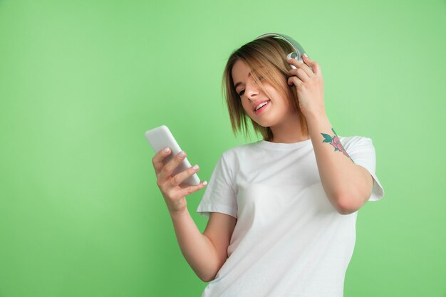 Ascoltare la musica. Il ritratto della giovane donna caucasica isolato sulla parete verde dello studio.