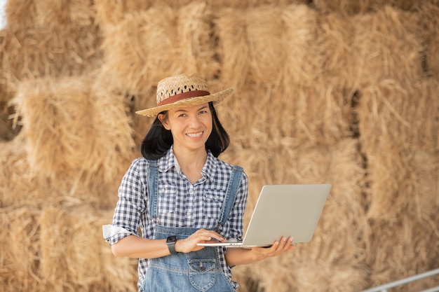 Asia giovane agricoltore femminile in cappello in piedi in campo e digitando sulla tastiera del computer portatile. donna con il computer portatile che supervisiona il lavoro sui terreni agricoli, concetto di ecologia, trasporti, aria pulita, cibo, prodotto biologico